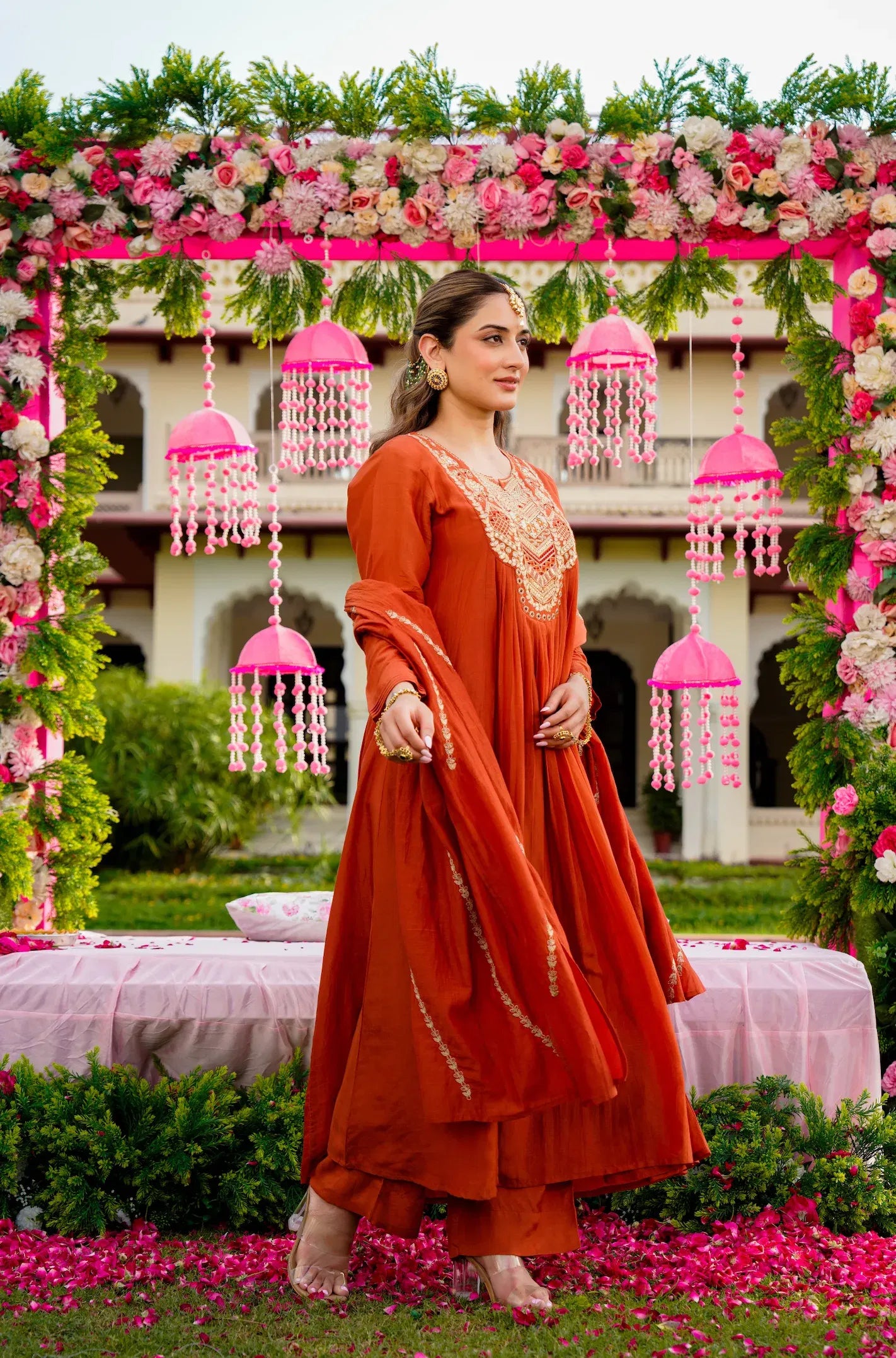 Woman in an orange traditional outfit standing in a decorated garden with pink floral arrangements.